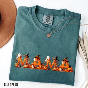 May include: Blue spruce Comfort Colors t-shirt with the word "MAMA" in orange letters decorated with Halloween-themed images. The design includes pumpkins, witch hats, ghosts, and black cats. A gold necklace is draped across the shirt.