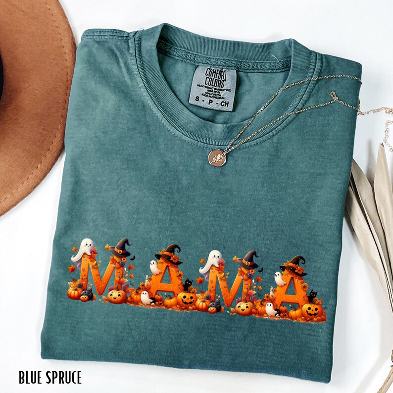 May include: Blue spruce Comfort Colors t-shirt with the word "MAMA" in orange letters decorated with Halloween-themed images. The design includes pumpkins, witch hats, ghosts, and black cats. A gold necklace is draped across the shirt.