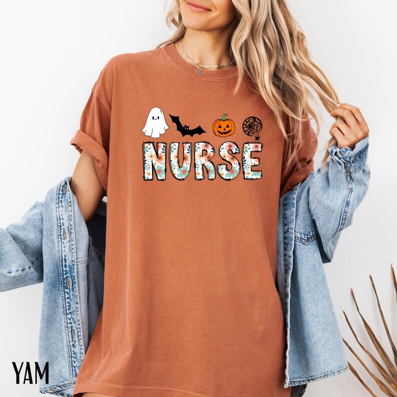 Puede incluir: Camiseta de color &oacute;xido con un dise&ntilde;o tem&aacute;tico de Halloween. La palabra "NURSE" est&aacute; impresa en letras grandes y florales, con un fantasma, un murci&eacute;lago, una calabaza y una telara&ntilde;a encima. La camiseta tiene las mangas enrolladas.