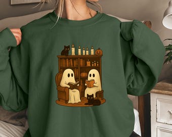 Puede incluir: Sudadera verde militar con una ilustración de Halloween. Dos figuras de fantasmas leen libros en sillones, con una estantería, velas y gatos negros. El texto "MILITARY GREEN" está en la parte inferior.