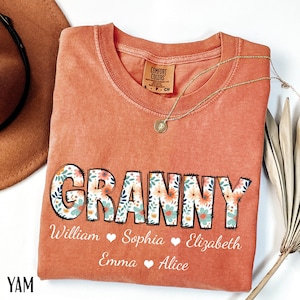 May include: A coral-colored Comfort Colors t-shirt with the word "GRANNY" in a floral design. Below the word are names: William, Sophia, Elizabeth, Emma, and Alice. A gold necklace and a brown hat are also visible.