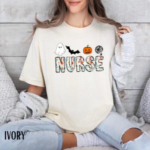 Puede incluir: Camiseta de color marfil con la palabra "NURSE" en un dise&ntilde;o floral, y gr&aacute;ficos tem&aacute;ticos de Halloween encima. Los gr&aacute;ficos incluyen un fantasma, un murci&eacute;lago, una calabaza y una telara&ntilde;a. La palabra "IVORY" est&aacute; impresa en la parte inferior.