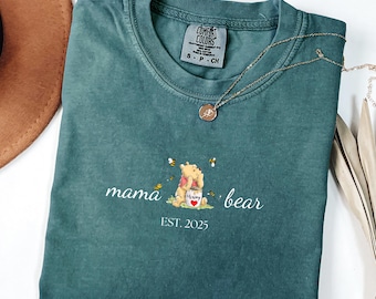 May include: A teal blue t-shirt with a Winnie the Pooh inspired design. The shirt features a small embroidered image of Winnie the Pooh holding a honey pot with the words "Mama Bear" and "EST. 2025" below the image.