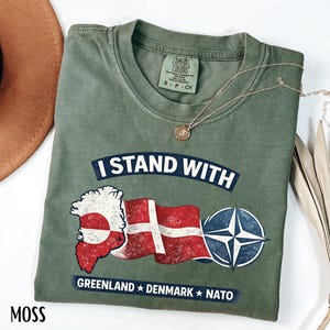 May include: Moss green t-shirt with the text "I STAND WITH" above a Greenland and Denmark flag graphic, and a NATO symbol. The text "GREENLAND * DENMARK * NATO" is below the graphic. A gold necklace is draped across the shirt.