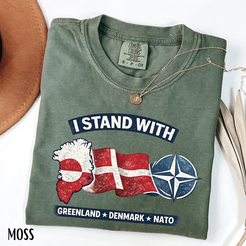 May include: Moss green t-shirt with the text "I STAND WITH" above a Greenland and Denmark flag graphic, and a NATO symbol. The text "GREENLAND * DENMARK * NATO" is below the graphic. A gold necklace is draped across the shirt.