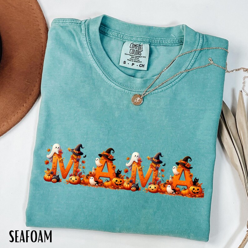 May include: Seafoam green t-shirt with the word "MAMA" in orange letters decorated with Halloween pumpkins, ghosts, and witch hats. A gold necklace is draped across the shirt. The shirt is a Comfort Colors brand.
