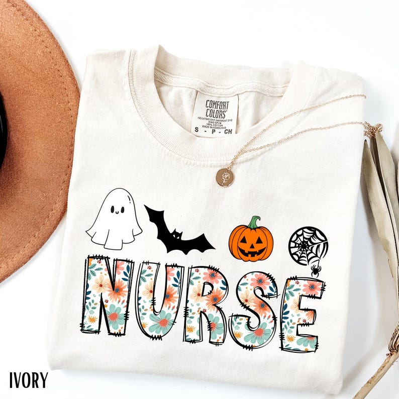 Puede incluir: Camiseta de color marfil con un dise&ntilde;o tem&aacute;tico de Halloween. La palabra "NURSE" est&aacute; escrita con letras florales, con un fantasma, un murci&eacute;lago, una calabaza y una telara&ntilde;a. Tambi&eacute;n se ve un collar dorado y un sombrero marr&oacute;n.