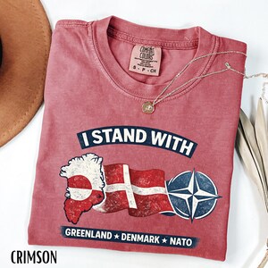 May include: Crimson-colored t-shirt with the text "I Stand With" above a graphic of Greenland, the Danish flag, and a NATO symbol. The text "Greenland Denmark NATO" is below the graphic. A silver necklace with a pendant is on the shirt.