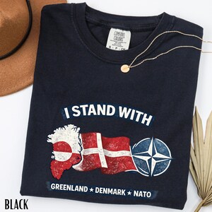 May include: Black Comfort Colors t-shirt with the text "I Stand With" above a graphic of the Greenland map, the Danish flag, and the NATO symbol. The words "Greenland Denmark NATO" are below the graphic.