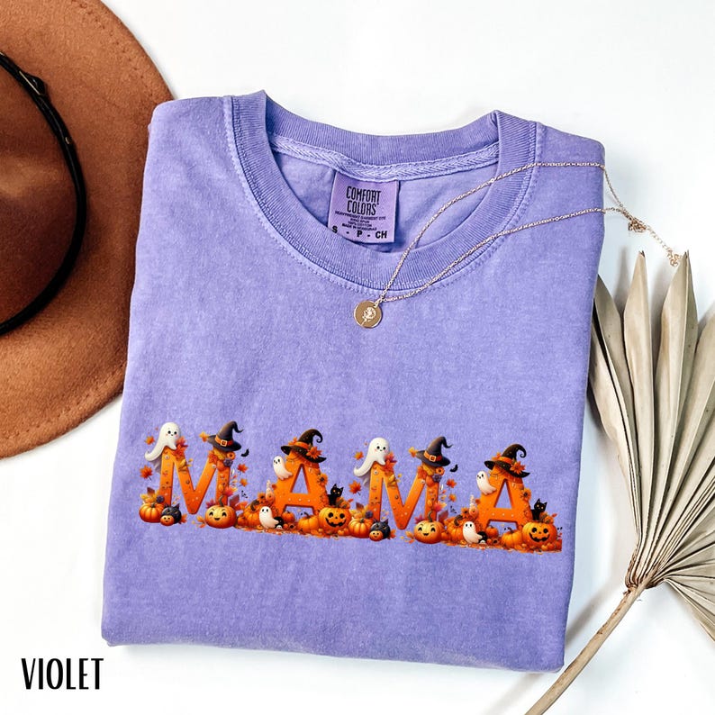 May include: A violet Comfort Colors t-shirt with the word "MAMA" in orange letters, decorated with Halloween-themed graphics. The design includes pumpkins, ghosts, witch hats, and black cats. A gold necklace is draped over the shirt.