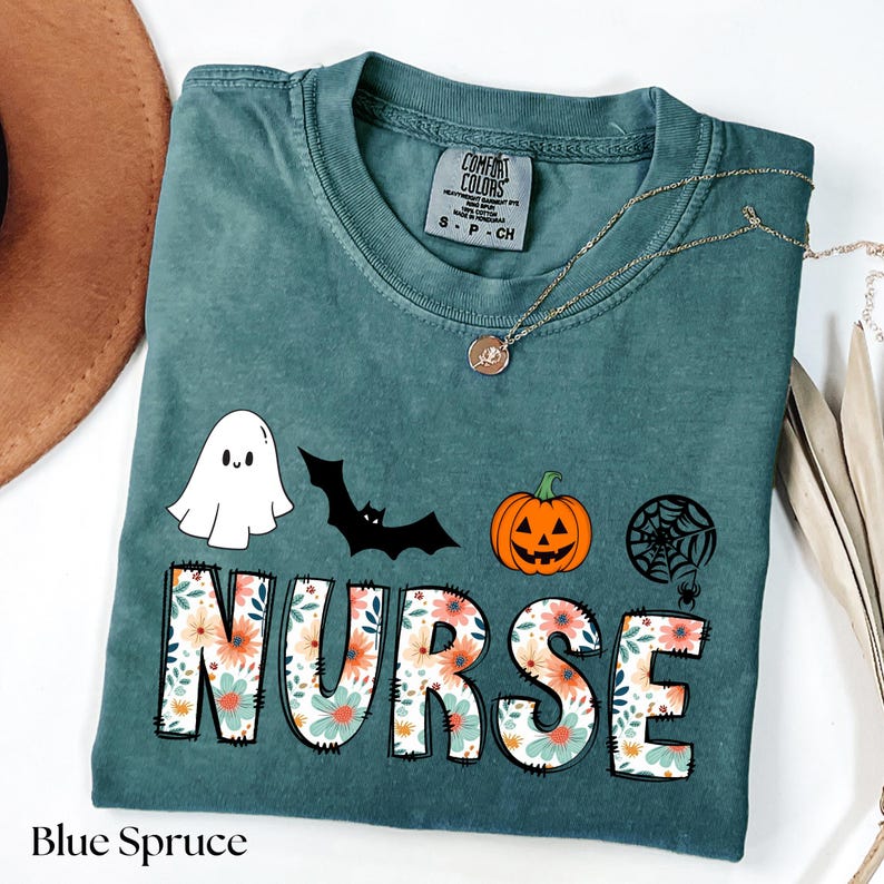 Puede incluir: Camiseta Comfort Colors azul abeto con dise&ntilde;o de Halloween. La palabra "NURSE" est&aacute; escrita en letras grandes con un estampado floral. La camiseta tambi&eacute;n presenta un fantasma, un murci&eacute;lago, una calabaza y una telara&ntilde;a.