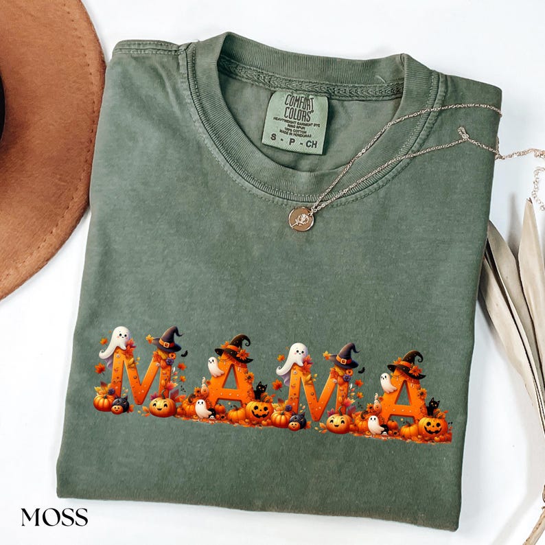 May include: Moss green Comfort Colors t-shirt with the word "MAMA" in orange letters, decorated with Halloween-themed graphics. The design includes pumpkins, ghosts, and witch hats. A silver necklace with a pendant is draped on the shirt.