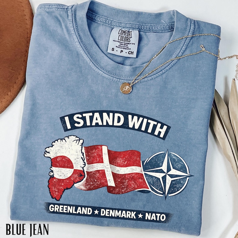 May include: A blue Comfort Colors t-shirt with the text "I STAND WITH" above a graphic of the Greenland and Danish flags, and a NATO symbol. The shirt also has the words "GREENLAND * DENMARK * NATO" printed on it. A gold necklace with a pendant is on the shirt.
