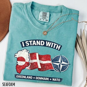 May include: Seafoam green t-shirt with the text "I STAND WITH" above a graphic of the Greenland and Danish flags, and a NATO symbol. The text "GREENLAND * DENMARK * NATO" is below the graphic. A gold necklace with a pendant rests on the shirt.