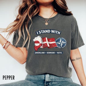 May include: Dark grey t-shirt with the text "I STAND WITH" above a graphic of the Greenland and Danish flags, and the NATO symbol. The text "GREENLAND * DENMARK * NATO" is below the graphic. The shirt is a comfortable, casual style.