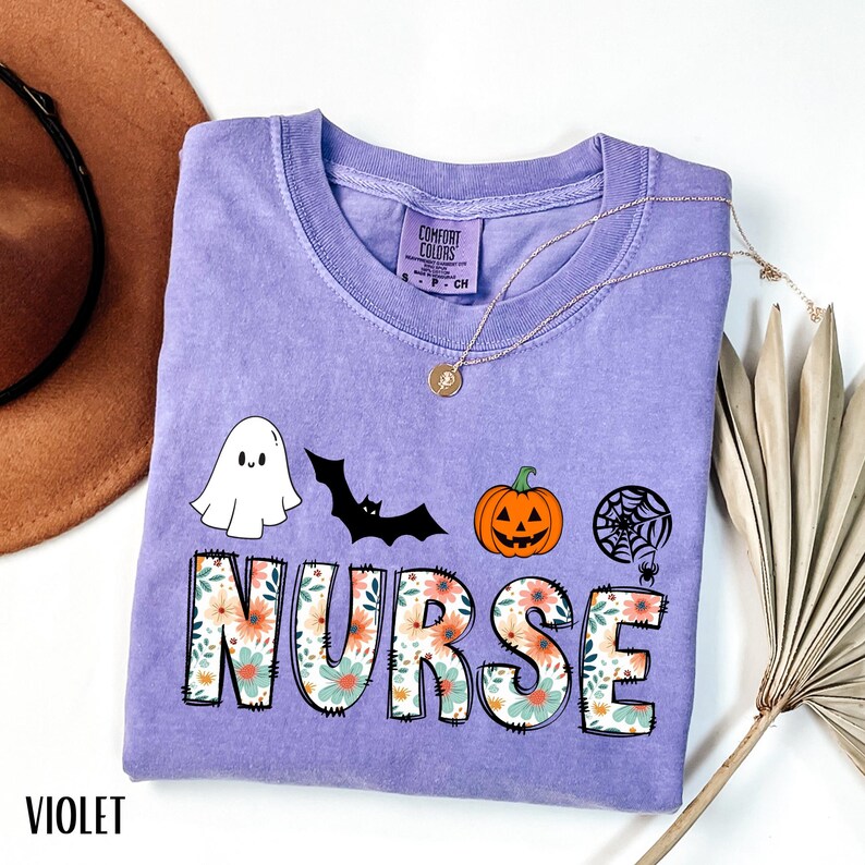 Puede incluir: Una camiseta violeta Comfort Colors con tem&aacute;tica de Halloween. La palabra "NURSE" est&aacute; impresa en letras grandes y florales, con un fantasma, un murci&eacute;lago, una calabaza y un gr&aacute;fico de telara&ntilde;a arriba. La palabra "VIOLET" est&aacute; impresa en la parte inferior.
