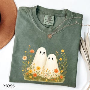 May include: Moss green Comfort Colors t-shirt featuring a graphic of two friendly ghosts surrounded by colorful flowers. The shirt has a crew neck and short sleeves. A silver necklace with a pendant is draped across the shirt.