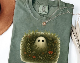 May include: A moss green t-shirt featuring a graphic of a friendly ghost surrounded by foliage and flowers. The shirt has a crew neck and short sleeves. A silver necklace with a small pendant is draped on the shirt. The word "MOSS" is at the bottom.