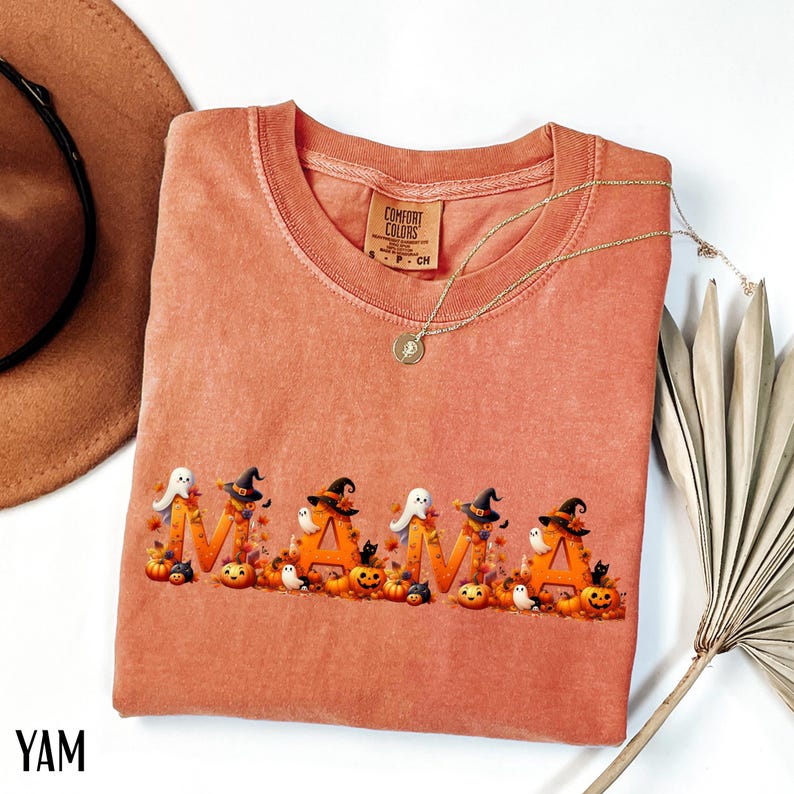 May include: A coral-colored Comfort Colors sweatshirt with the word "MAMA" in orange letters, decorated with Halloween-themed graphics. The design includes pumpkins, ghosts, witch hats, and fall leaves. A gold necklace and a brown hat are also visible.