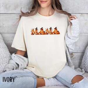 May include: Ivory-colored t-shirt with the word "MAMA" in orange letters, each decorated with a witch hat and pumpkins. The shirt is a comfortable fit, perfect for Halloween or fall. The word "IVORY" is printed at the bottom.