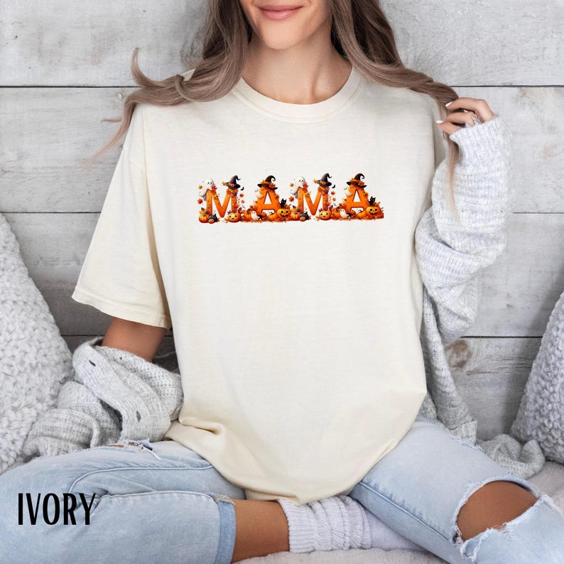 May include: Ivory-colored t-shirt with the word "MAMA" in orange letters, each decorated with a witch hat and pumpkins. The shirt is a comfortable fit, perfect for Halloween or fall. The word "IVORY" is printed at the bottom.