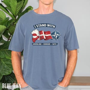 May include: Blue jean colored t-shirt with the text "I Stand With" above a graphic of Greenland, the Danish flag, and the NATO symbol. Below the graphic are the words "Greenland Denmark NATO".