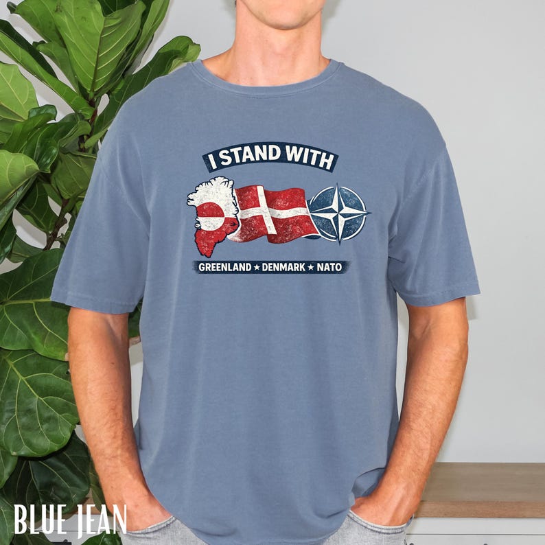 May include: Blue jean colored t-shirt with the text "I Stand With" above a graphic of Greenland, the Danish flag, and the NATO symbol. Below the graphic are the words "Greenland Denmark NATO".