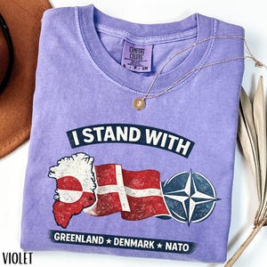 May include: Violet-colored t-shirt with the text "I STAND WITH" above a graphic of the Greenland and Denmark flags, and a NATO symbol. The text "GREENLAND * DENMARK * NATO" is below the graphic. A gold necklace with a pendant is on the shirt.