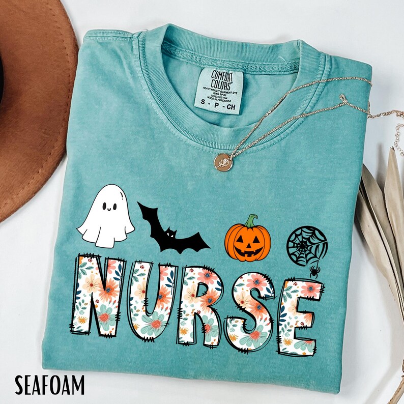 Puede incluir: Camiseta verde mar con la palabra "NURSE" en letras florales grandes. La camiseta presenta un fantasma, un murci&eacute;lago, una calabaza de Halloween y un gr&aacute;fico de telara&ntilde;a. Un collar dorado est&aacute; drapeado sobre la camisa.