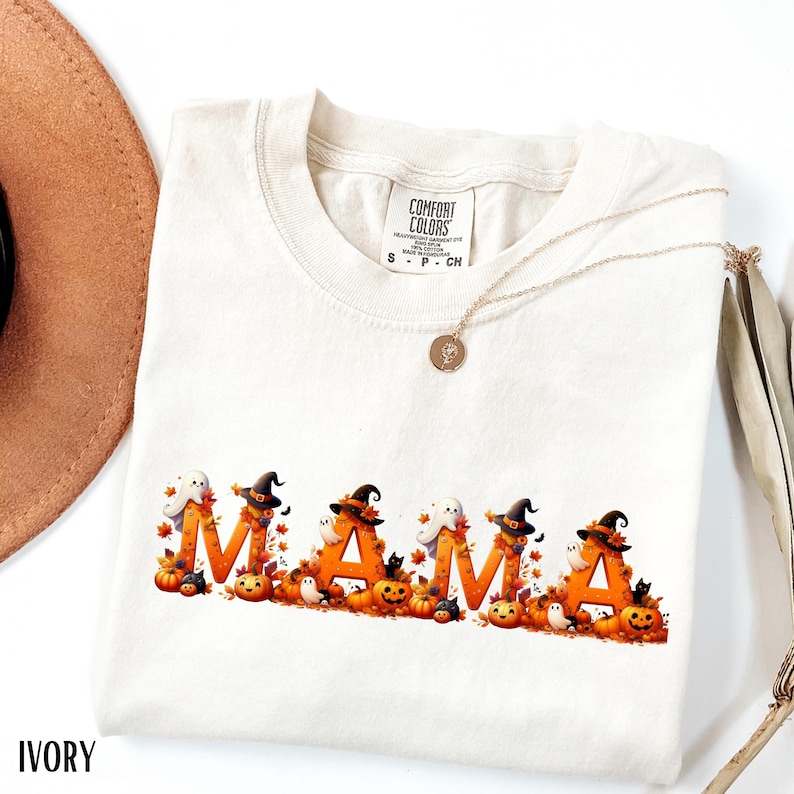 May include: Ivory-colored t-shirt with the word "MAMA" in orange letters, decorated with Halloween-themed graphics. The design includes pumpkins, witch hats, ghosts, and fall leaves. A gold necklace and a brown hat are also visible.