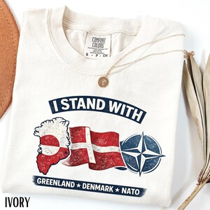 May include: Ivory-colored t-shirt with a graphic that reads "I Stand With" above a Greenland map, Danish flag, and NATO symbol. The text "Greenland Denmark NATO" is below the graphics. A gold necklace with a pendant is draped on the shirt.