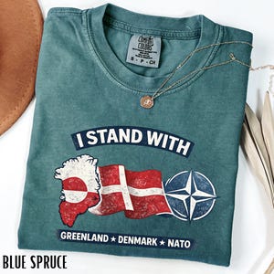 May include: Blue spruce t-shirt with the text "I Stand With" above a graphic of the Greenland map, the Danish flag, and a NATO symbol. The shirt also has the text "Greenland Denmark NATO" below the graphic.