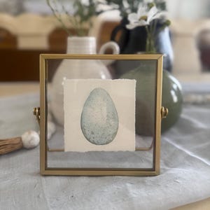 May include: A framed watercolor painting of a speckled blue egg. The egg is centered on a textured white paper, encased in a gold-colored metal frame. The background includes vases with white flowers and a light blue fabric.