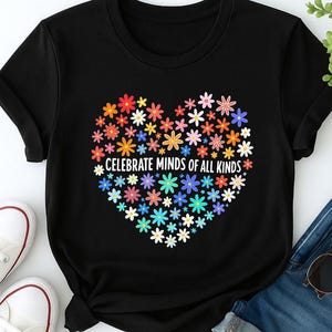 Peut inclure: T-shirt noir avec un motif floral en forme de cœur composé de nombreuses petites fleurs colorées. Le texte "CELEBRATE MINDS OF ALL KINDS" est imprimé en blanc. Le t-shirt est associé à un jean bleu et des baskets blanches.