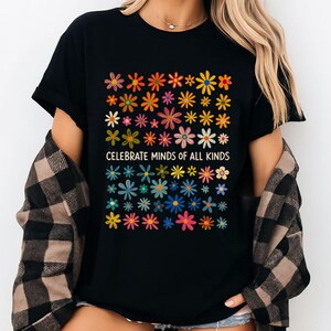 May include: Black t-shirt with a colorful floral design and the text "CELEBRATE MINDS OF ALL KINDS". The flowers are in various colors, including red, orange, yellow, green, blue, and pink. The shirt is worn with a plaid shirt.