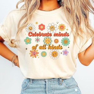 Könnte beinhalten: Cremefarbenes T-Shirt mit einem farbenfrohen Blumendesign und lächelnden Gesichtern. Der Text "Celebrate minds of all kinds" ist über den Blumen aufgedruckt. Das Shirt hat kurze Ärmel und eine lockere Passform.