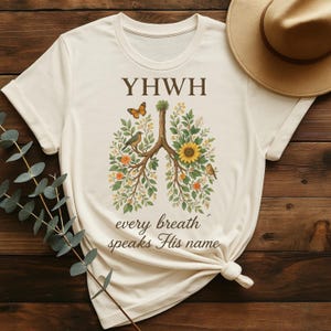 Può includere: Una maglietta beige con un disegno floreale di polmoni con la parola "YHWH" sopra il disegno. Il testo "every breath speaks His name" è sotto il disegno.