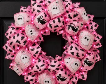 Puede incluir: Una corona decorativa redonda con lazos de cinta a rayas rosas y blancas, intercalados con patrones de murciélagos negros, ilustraciones de fantasmas blancos y siluetas de gatos negros. La corona está iluminada por pequeñas luces LED rosas.