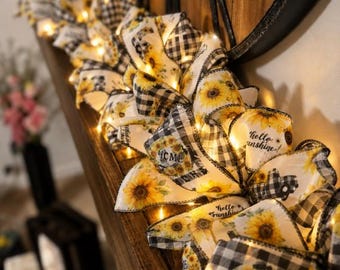 Könnte beinhalten: Eine dekorative Girlande aus Drahtband mit Sonnenblumenmotiven, schwarz-weißem Karomuster und dem Schriftzug "HOME" sowie "hello sunshine!", durchzogen von warmweißen Lichterketten in den verschiedenen Stoffschleifen.