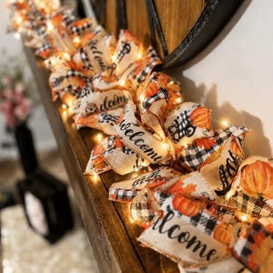 K&ouml;nnte beinhalten: Eine dekorative Girlande aus beigefarbenen Stoffb&auml;ndern mit schwarz-wei&szlig;em Karomuster, orangefarbenen K&uuml;rbisillustrationen und dem in schwarzer Schrift gedruckten Wort Welcome. Die Girlande wird von kleinen, warmwei&szlig;en Lichterketten beleuchtet.