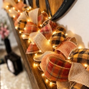 May include: A decorative garland featuring loops of hessian ribbon interspersed with orange, red, and black plaid fabric ribbons. The garland is illuminated by a string of small, warm white LED fairy lights woven throughout the textured fabric loops.