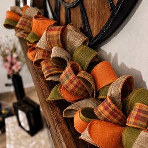 Puede incluir: Una guirnalda decorativa compuesta por bucles de cinta con alambre en texturas y colores alternos. La cinta presenta naranja sólido, verde oliva sólido y una tela estilo arpillera color canela, junto a un patrón de cuadros en tonos naranja, rojo y marrón.