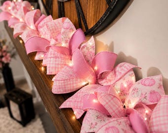 Könnte beinhalten: Eine dekorative Girlande aus rosa Stoffbändern mit verschiedenen Mustern wie Vichy-Karo, einfarbigem Rosa und Kürbisdruck, durchsetzt mit kleinen warmweißen LED-Lichterketten, die in Schleifen auf einer Holzoberfläche angeordnet sind.