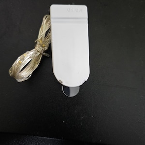May include: A white battery pack with an "ON OFF" switch, connected to a string of small, warm white LED lights. The lights are coiled next to the battery pack. The battery pack is on a black surface.