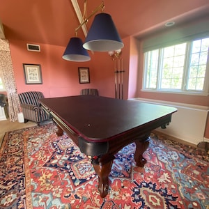 May include: A dark wood pool table with a red felt top and a dark brown leather trim. The table is in a room with a red wall, a window with white trim, and a patterned rug.