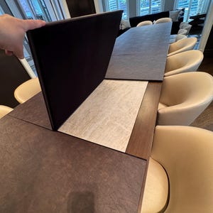 May include: A large, rectangular dining table with a dark brown surface and a light-colored center section. The table is surrounded by cream-colored chairs. A dark panel is being lifted, revealing the table's construction.