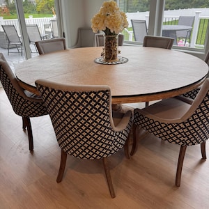 May include: A round dining table with a light wood finish, surrounded by six upholstered chairs. The chairs have patterned backs in black and white. A vase of white flowers sits on the table's center. The table is approximately 60 inches in diameter.