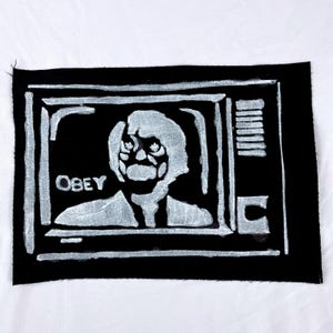May include: Black patch with a white graphic of a face inside a television screen. The word "OBEY" is printed in white at the bottom of the screen. The patch is on a white background.