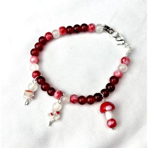 Puede incluir: Una pulsera de cuentas rojas y blancas con dijes de setas. La pulsera tiene un cierre plateado y está hecha con pequeñas cuentas redondas. Los dijes incluyen una seta roja y blanca y otros dijes pequeños.