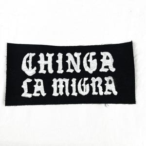 May include: Black rectangular patch with the words "CHINGA LA MIGRA" embroidered in white, stylized lettering. The patch is made of a felt-like material.